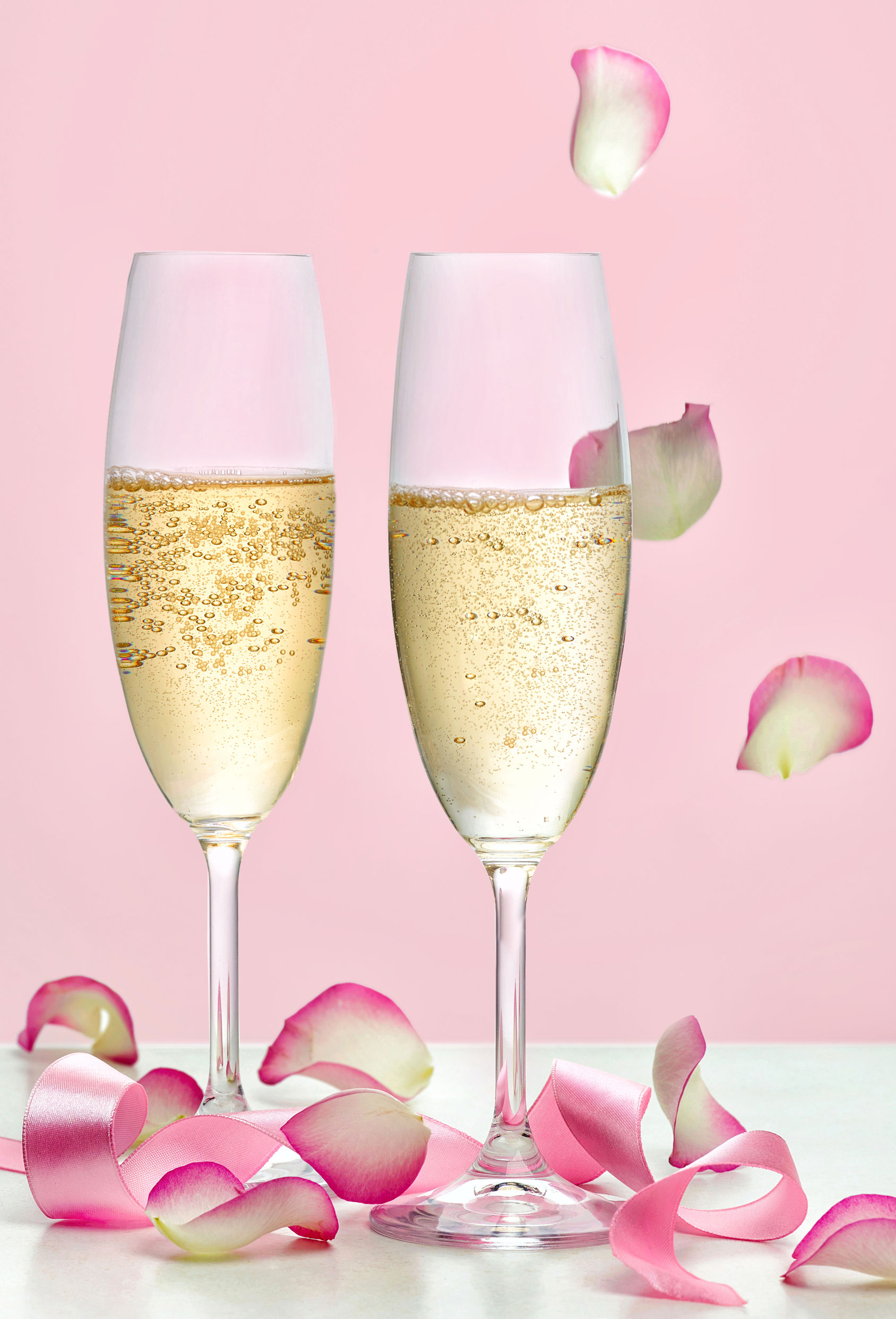 Two glasses of champagne with rose petals against pink backdrop
